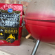 Bag of charcoal briquets beside red camping grill whose color has singed off the top